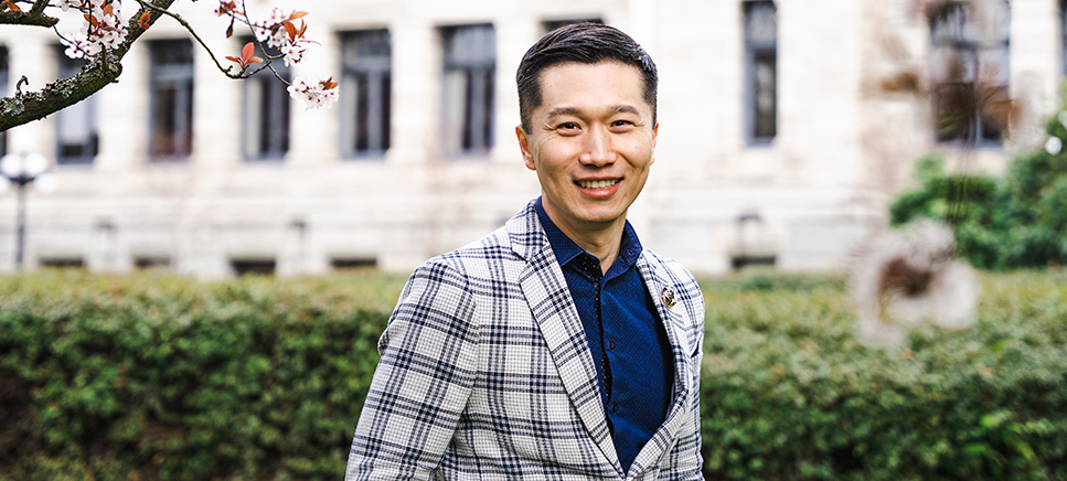 Person in a grey plaid blazer and blue shirt smiling outdoors, with greenery and a building in the background 